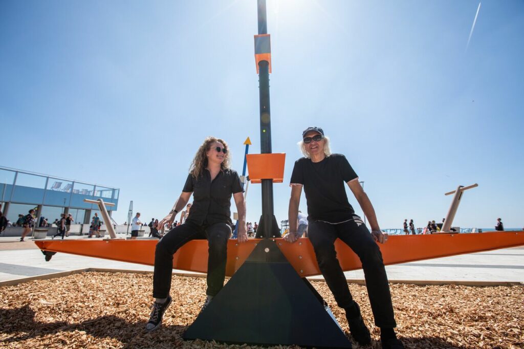 A man and a woman sit on a seesaw artwork outside in the sun. They both wear black.